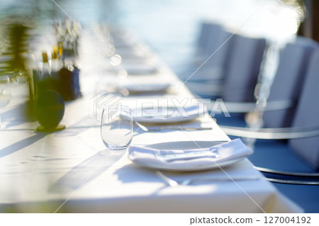 A row of empty plates on a long table with a white tablecloth. A table in a Mediterranean restaurant ready to serve a large group of visitors or tourists. 127004192