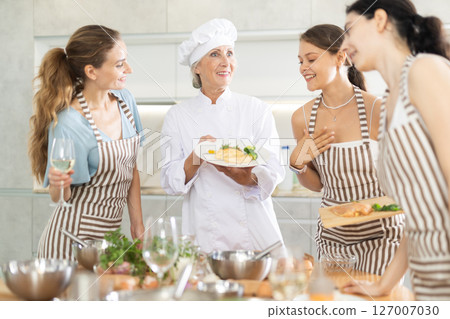 Senior woman host of cooking courses shows visitors of workshop cooked chicken under creamy sauce 127007030