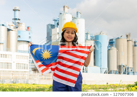 Positive girl in overalls with Malaysia flag in her hands against the background of chimneys of modern factory 127007306