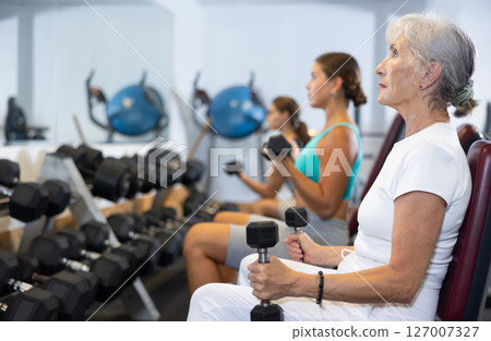Mature sportswoman doing exercises with a barbell and dumbbells in gym Mature sportswoman doing exercises with a barbell and dumbbells in gym 127007327