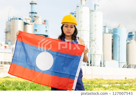 Sad young woman in helmet holding flag of Laos against background of factory 127007543