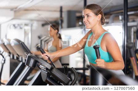 Slender athletic girl during cardio exercises on elliptical simulator in fitness center. Slender athletic girl during cardio exercises on elliptical simulator in fitness center. 127007560