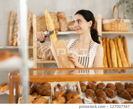Young woman works as salesperson in bakery and packs baguettes in paper bag for customer 127007567