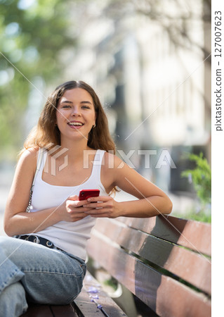 Cheerful young girl messaging on smartphone on bench 127007623
