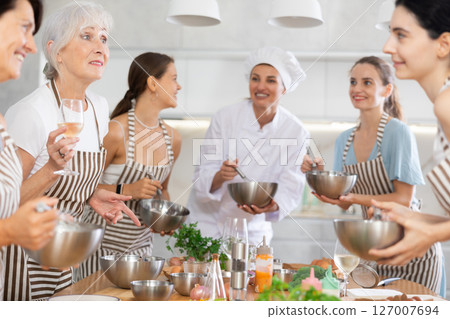 Aged woman holding wine and talking to other cooking course members whipping souce in bowls 127007694
