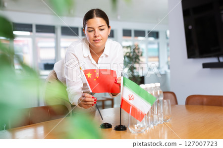 A woman in a white blouse with friendly smile arrangs flags of China and Iran for meeting. A woman in a white blouse with friendly smile arrangs flags of China and Iran for meeting. 127007723
