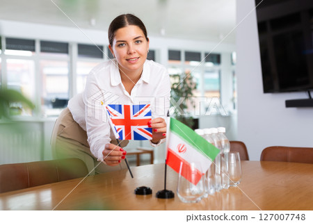 A woman in a white blouse with friendly smile arrangs flags of Britain and Iran for meeting. 127007748