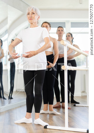 Women stand in fifth position near ballet barre during group training in dance studio Women stand in fifth position near ballet barre during group training in dance studio 127007770