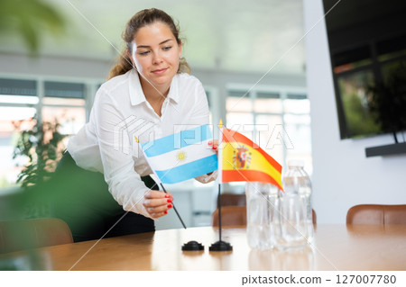 Positive young woman putting little flag of Argentina on table next to the flag of Spain 127007780