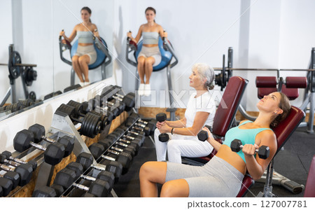 Slender girl sit and trains muscles of arms and back using dumb-bell shaped figure weighting. 127007781