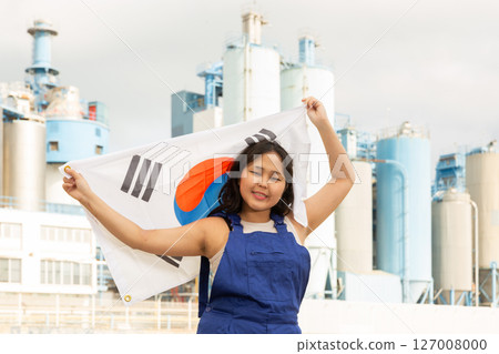 Portrait of teenage girl in blue overalls with flag of South Korea in her hands against the backdrop of modern metallurgical plant 127008000