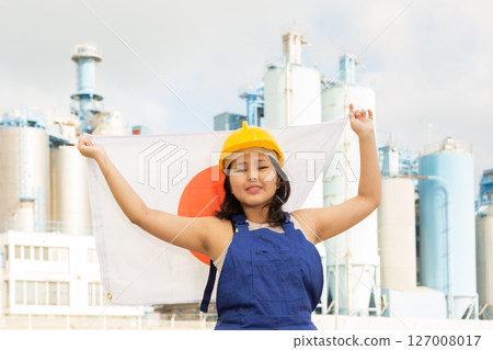 Happy young woman in helmet holding flag of Japan against background of factory Happy young woman in helmet holding flag of Japan against background of factory 127008017