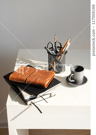 Minimalist Workspace Featuring Desk with Various Notebooks, Coffee Cup and Holder With Pens and Scissors. 127008166