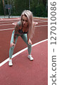 Young blonde woman in sportswear doing stretching exercises standing on basketball court in the morning. Woman feels pleasantly tired after intense daily workout program. Genuine candid photo 127010086
