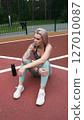 The concept of energy, vitality, well-being. A toned, beautiful blonde in sports clothes is relaxing after a workout outdoors. She's sitting on a sports basketball court 127010087