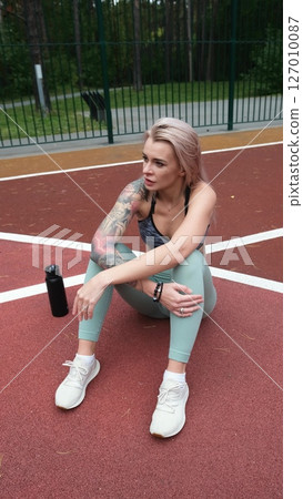 The concept of energy, vitality, well-being. A toned, beautiful blonde in sports clothes is relaxing after a workout outdoors. She's sitting on a sports basketball court 127010087