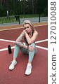 The concept of energy, vitality, well-being. A toned, beautiful blonde in sports clothes is relaxing after a workout outdoors. She's sitting on a sports basketball court 127010088