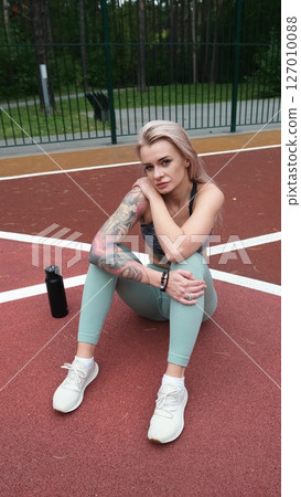 The concept of energy, vitality, well-being. A toned, beautiful blonde in sports clothes is relaxing after a workout outdoors. She's sitting on a sports basketball court 127010088
