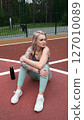 The concept of energy, vitality, well-being. A toned, beautiful blonde in sports clothes is relaxing after a workout outdoors. She's sitting on a sports basketball court 127010089