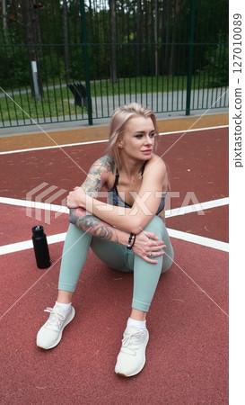 The concept of energy, vitality, well-being. A toned, beautiful blonde in sports clothes is relaxing after a workout outdoors. She's sitting on a sports basketball court 127010089