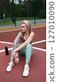 The concept of energy, vitality, well-being. A toned, beautiful blonde in sports clothes is relaxing after a workout outdoors. She's sitting on a sports basketball court 127010090
