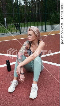 The concept of energy, vitality, well-being. A toned, beautiful blonde in sports clothes is relaxing after a workout outdoors. She's sitting on a sports basketball court 127010090