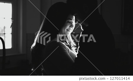 Married couple hugging in the morning in the kitchen. Man and woman in love hugging in the rays of the morning sun. Monochrome image.  127011414