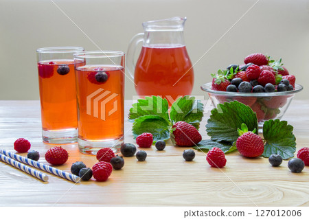Left two glasses of berries compote, near bowl with berries, carafe compote on a light wooden background scattered berries and some empty space . Fresh berries compote and empty space. Horizontal. Left two glasses of berries compote, near bowl with berries, carafe compote on a light wooden background scattered berries and some empty space . Fresh berries compote and empty space. Horizontal. 127012006