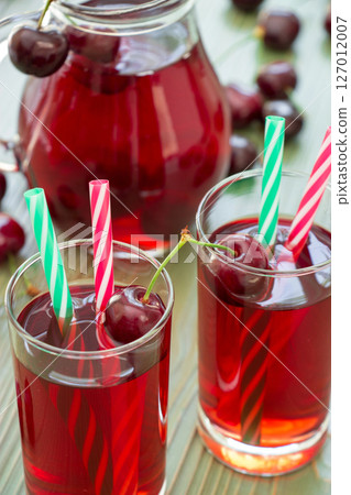 Two glasses of cherry juice with straws in the foreground and in the background a jar full of cherry juice. 2 cherry juices. Vertical. Close-up. Daylight. 127012007