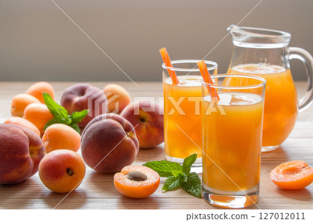Apricot and peach juice in glasses with ice near the jar of juice and scattered apricots and peaches, mint on a light wooden background. Apricot and peach juice with ice. Horizontal. Daylight. Close. Apricot and peach juice in glasses with ice near the jar of juice and scattered apricots and peaches, mint on a light wooden background. Apricot and peach juice with ice. Horizontal. Daylight. Close. 127012011