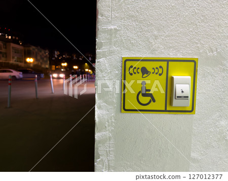 Accessible call button for people with disabilities mounted on textured wall in urban area at night. Clear signage with tactile surface and visual symbol for inclusive design 127012377