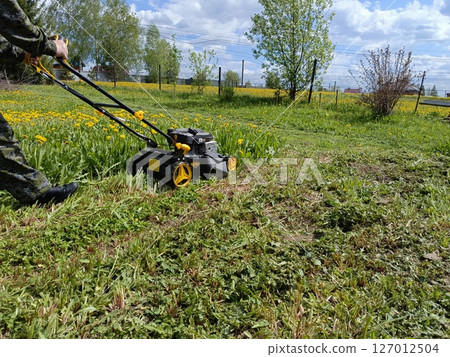 Person in Camouflage Pants Diligently Operating Lawnmower in Sunny Dandelion Field, Performing Yard Maintenance, Against Chain Link Fence Person in Camouflage Pants Diligently Operating Lawnmower in Sunny Dandelion Field, Performing Yard Maintenance, Against Chain Link Fence 127012504