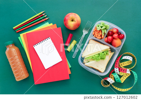 Lunch box includes sandwich with lettuce, fruits, juice bottle, colored notebooks and pencils placed on green surface ready for school day activities. 127012602