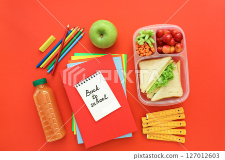 School supplies and lunch box with sandwiches, pieces of cucumbers and carrots, apricot, cherries, apple, bottle of juice on red background, back to school concept. Horizontal. Top view. School supplies and lunch box with sandwiches, pieces of cucumbers and carrots, apricot, cherries, apple, bottle of juice on red background, back to school concept. Horizontal. Top view. 127012603