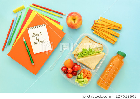Lunchbox features sandwiches, fresh vegetables, fruits, along with stationery items like notebooks, pencils, and rulers on bright blue surface, preparing for school day. Lunchbox features sandwiches, fresh vegetables, fruits, along with stationery items like notebooks, pencils, and rulers on bright blue surface, preparing for school day. 127012605