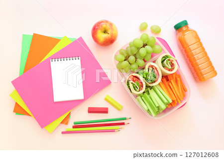 School supplies and lunch box with tasty rolls, cucumbers, carrots, grapes, apple, bottle of juice on light pink background, back to school concept. Horizontal. Top view. 127012608