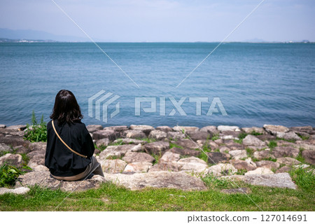 Back view of a woman sitting on the shore of Lake Biwa and looking at the lake 127014691