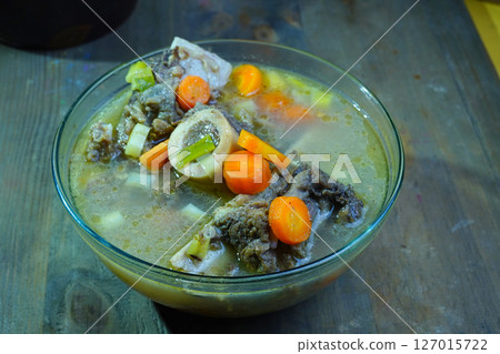 A close-up of a hearty bone marrow soup, featuring tender meat, vibrant carrots, and green vegetables in a clear glass bowl on a rustic wooden table. A close-up of a hearty bone marrow soup, featuring tender meat, vibrant carrots, and green vegetables in a clear glass bowl on a rustic wooden table. 127015722