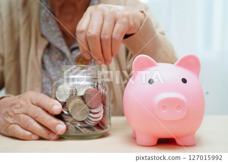 Retired elderly woman counting coins money with piggy bank and worry about monthly expenses and treatment fee payment. Retired elderly woman counting coins money with piggy bank and worry about monthly expenses and treatment fee payment. 127015992
