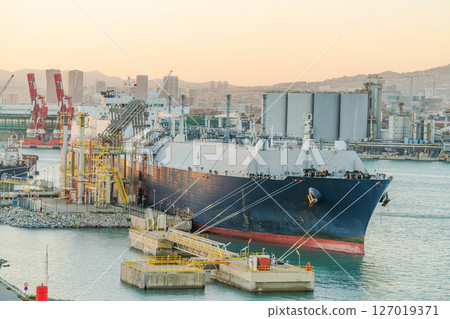 Large LNG (liquefied natural gas) tanker moored at an industrial port terminal with yellow pipelines, storage tanks, and cranes 127019371