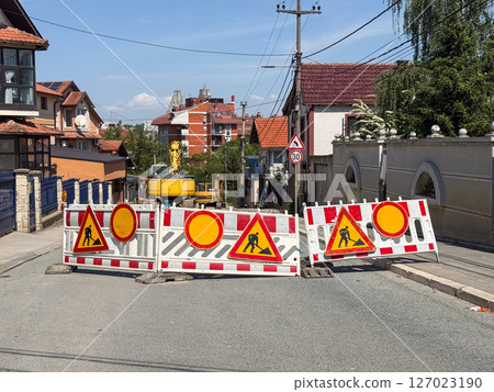 Street closed for construction work. Traffic barriers with warning signs and excavator visible in residential area. Street closed for construction work. Traffic barriers with warning signs and excavator visible in residential area. 127023190
