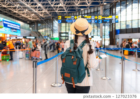 Back view of Caucasian woman in a straw hat, wearing backpack, walks through the airport. Traveling by plane, check-in for a flight. Concept of journey 127023376