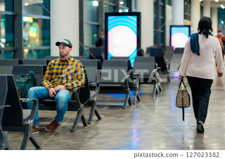 Wide shot of adult Caucasian man in cap and jacket using smartphone in airport waiting area sitting in chair. Concept of boarding at terminal before flight 127023382