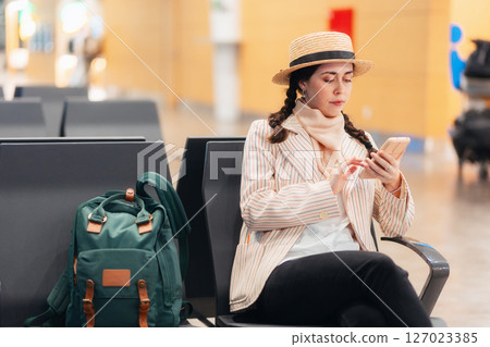 Mid shot of young Caucasian elegant woman is sitting in lounge chair at airport waiting room and using smartphone. Concept of travel and trip 127023385