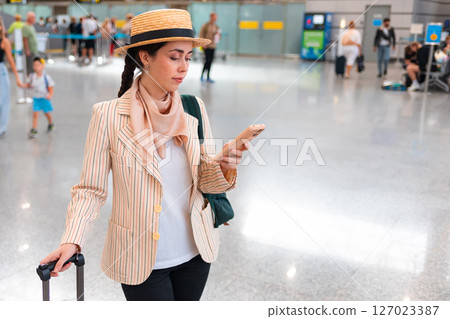 Mid shot of young pretty Caucasian woman in an elegant suit uses a smartphone and holds luggage. Person in airport. Copy space. Concept of trip and travel 127023387