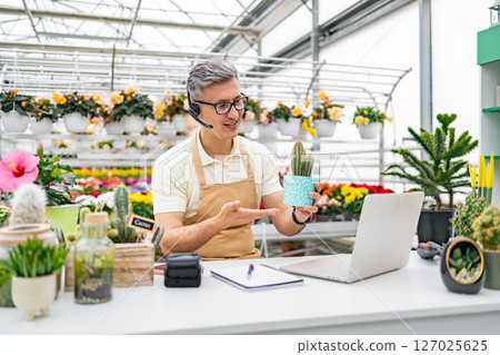 A Caucasian florist conducts a video call while showcasing a cactus for sale, highlighting online sales through a laptop. A Caucasian florist conducts a video call while showcasing a cactus for sale, highlighting online sales through a laptop. 127025625