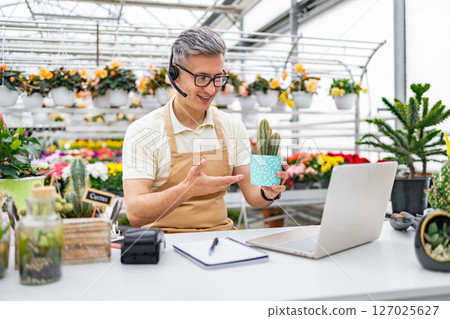 A florist on a video call, showing a cactus to a customer. The shop has a large selection of flowers. 127025627