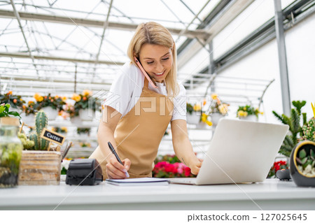 A Caucasian woman working in a florist shop, taking a call and using a laptop while writing. A Caucasian woman working in a florist shop, taking a call and using a laptop while writing. 127025645