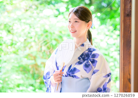 身著浴衣、手持扇子、面帶溫柔表情的女子，背景為綠色植物 127026859