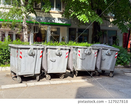 Row of metal garbage bins on city street. Industrial waste containers with wheels placed near sidewalk and greenery. 127027818
